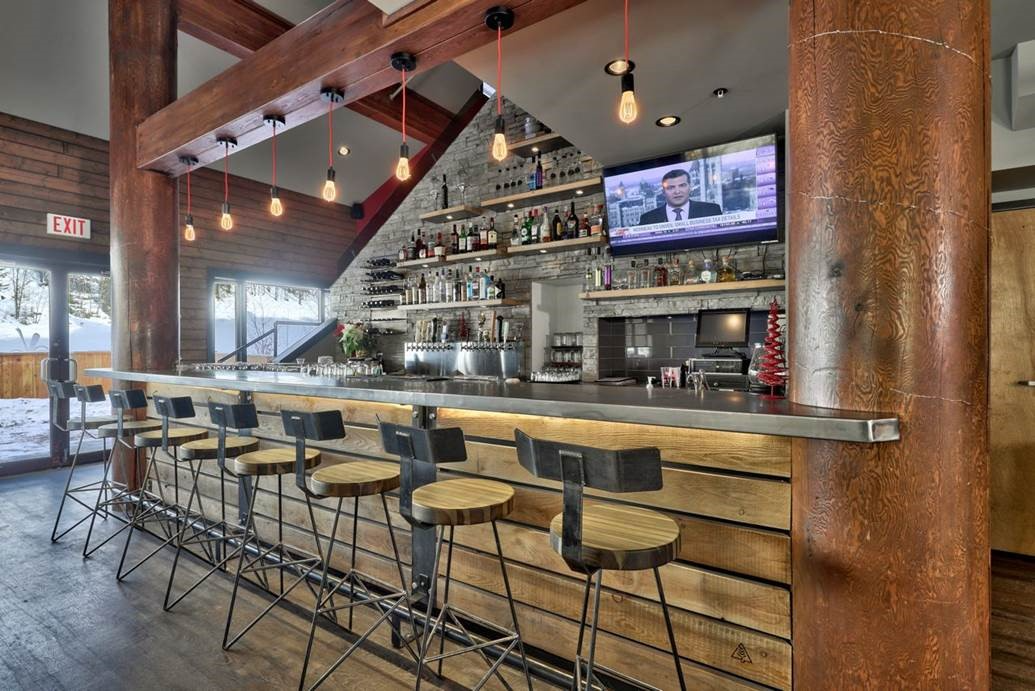 A modern bar features wooden accents, illuminated by hanging bulbs and a television display