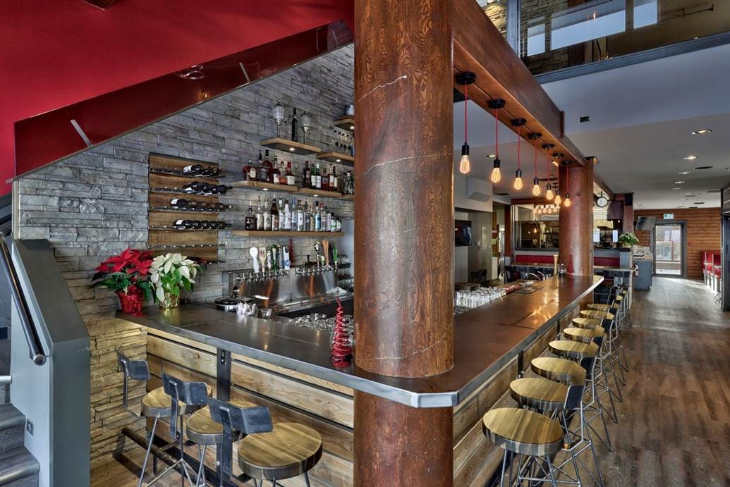A stylish bar features wooden beams, shelves of bottles, and modern stools