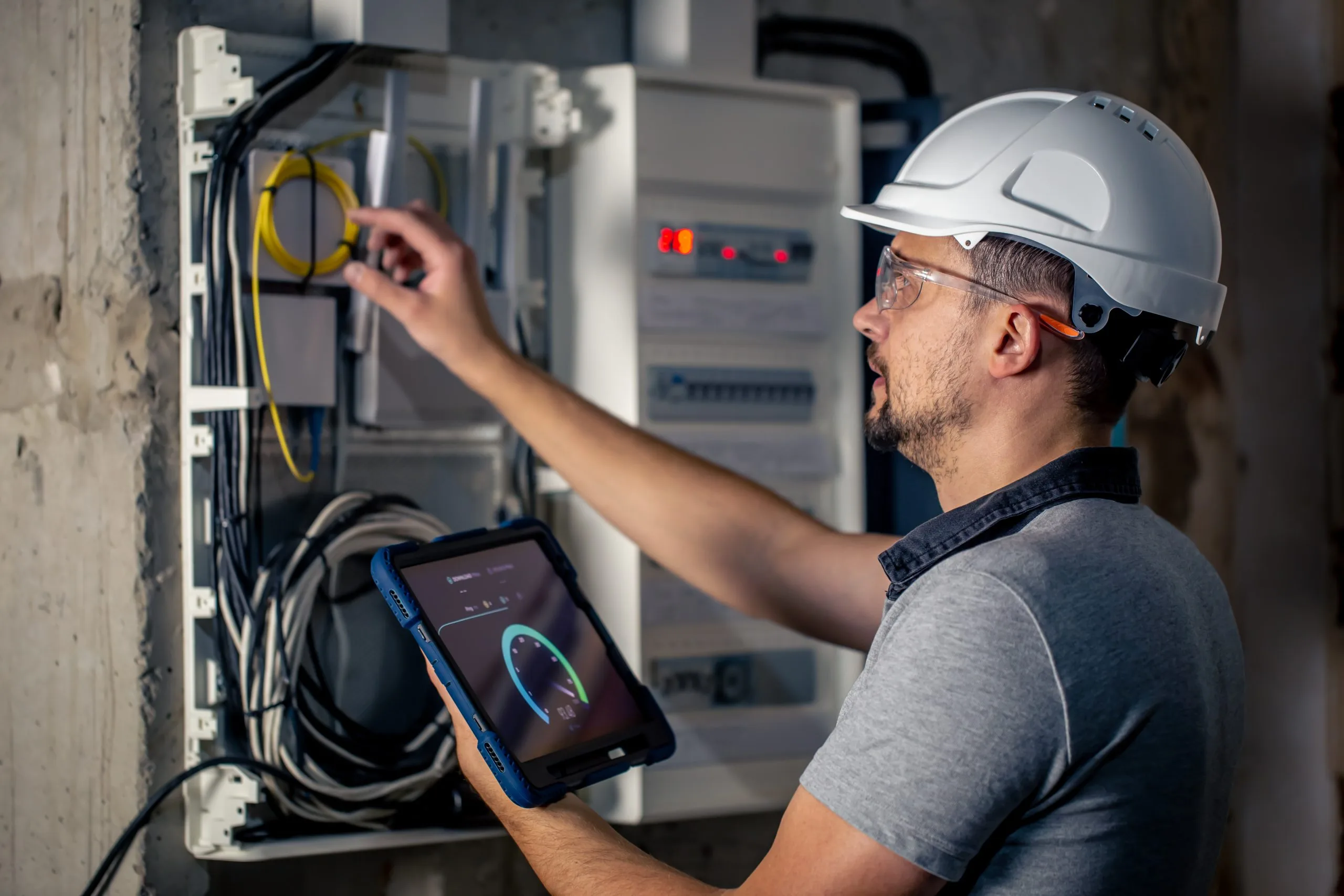 A technician in a hard hat is checking electrical connections while using a tablet