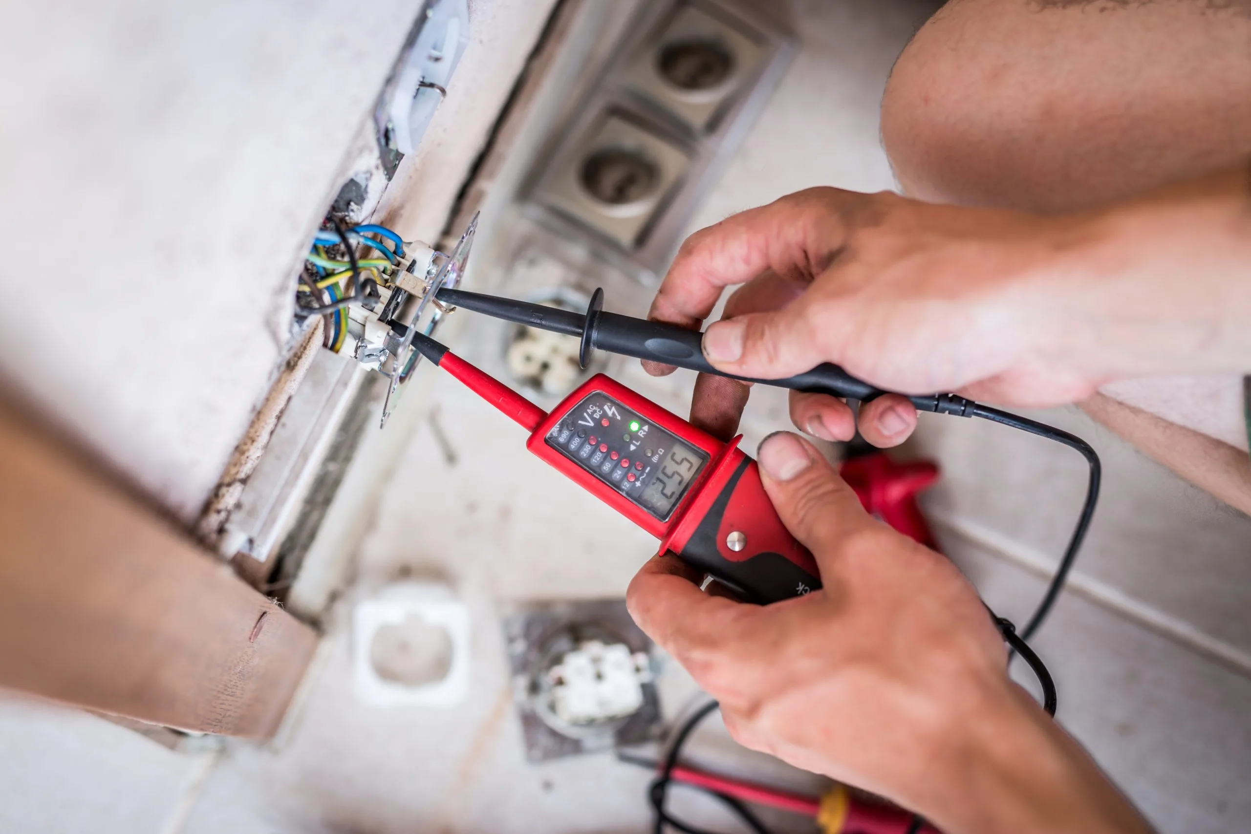 Hands hold a multimeter while testing electrical wiring at a wall outlet