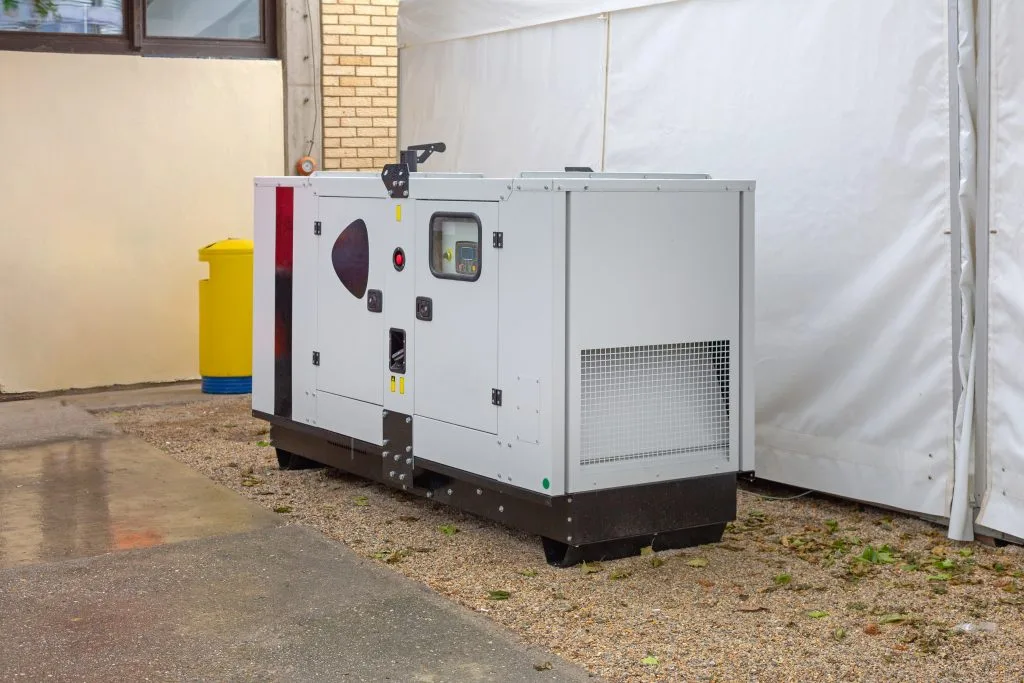 A generator sits quietly beside a building, partially sheltered by a tent