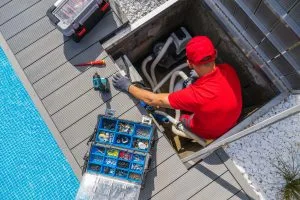 A worker is repairing pool equipment, surrounded by tools and accessories